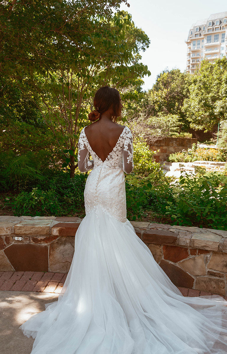 Woman in Style 7491 by Essense of Australia - Long Sleeve Lace Fit-and-Flare Wedding Dress with Train with a low back, standing in a garden setting.
