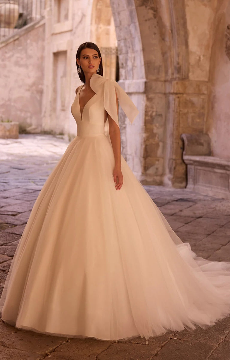 Woman in Yudita by Veni Infantino Bridal - Princess A-Line Ball Gown with Statement Bow Detail standing in an outdoor stone setting