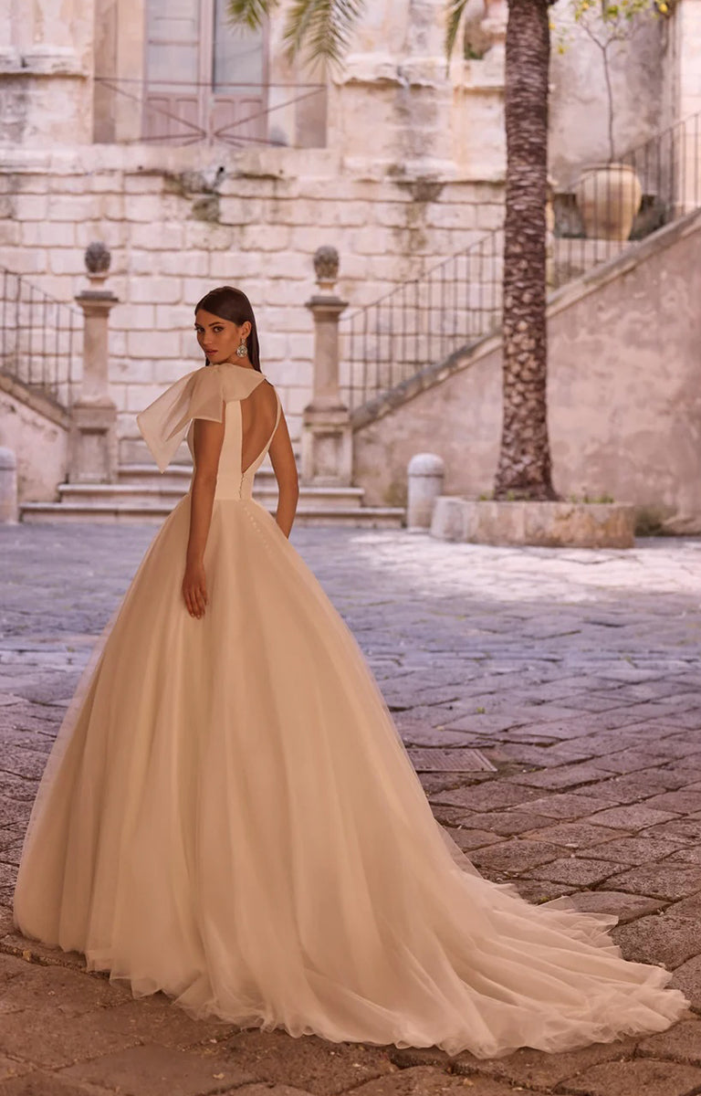 Woman in Yudita by Veni Infantino Bridal - Princess A-Line Ball Gown with Statement Bow Detail standing in an outdoor setting with stone steps and a palm tree.