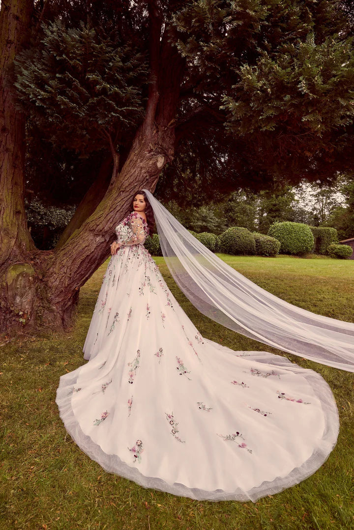 Woman in a white wedding dress with a long train standing under a tree in a garden.
