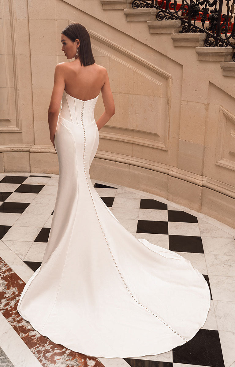 Woman in a white strapless gown standing on a checkered floor with decorative wall and staircase.