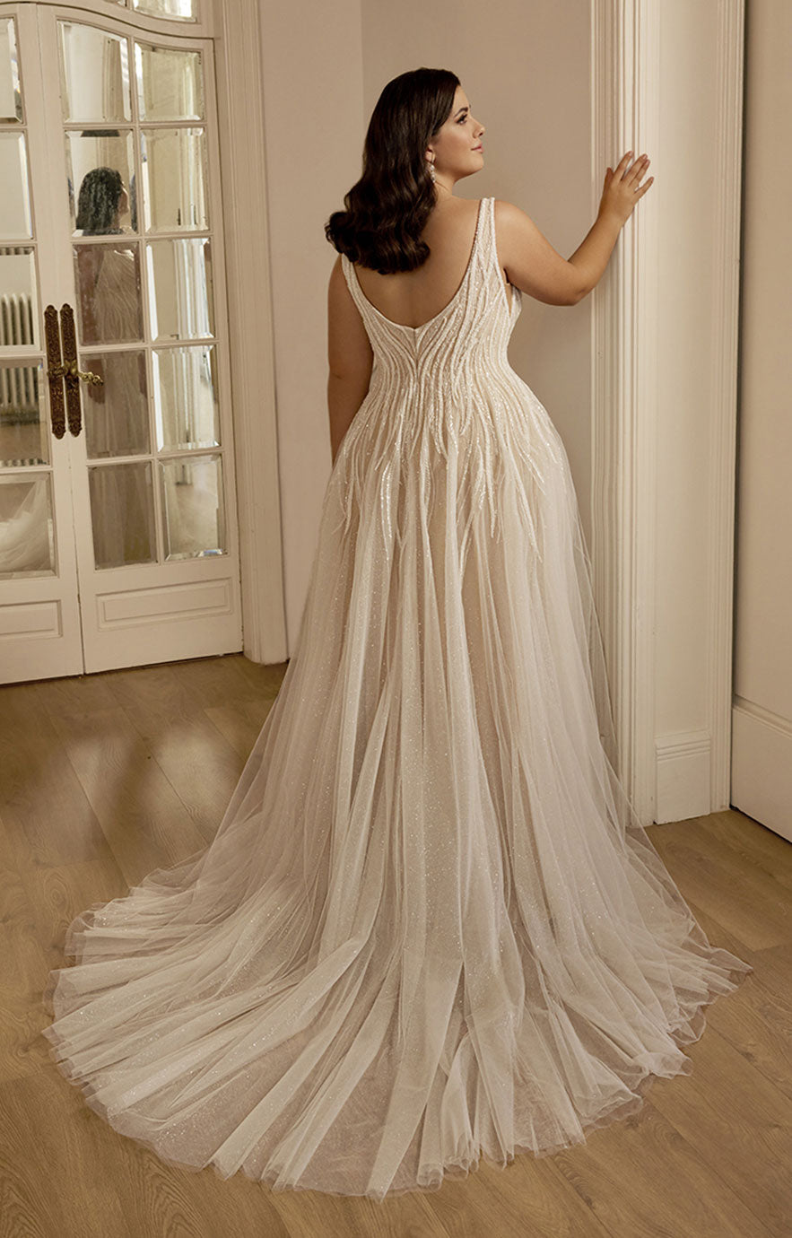 Woman in a flowing white dress standing in a room with a mirror and door.