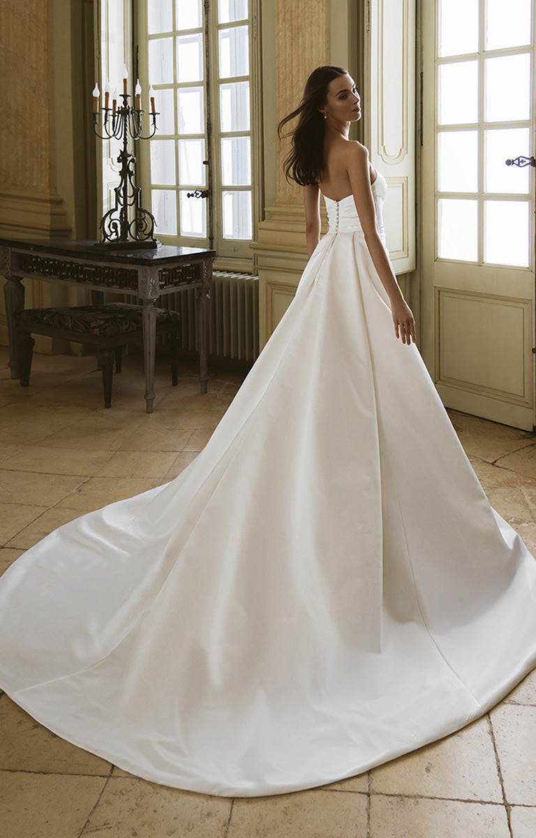 Woman in a white wedding dress standing in a room with large windows and a wooden floor.