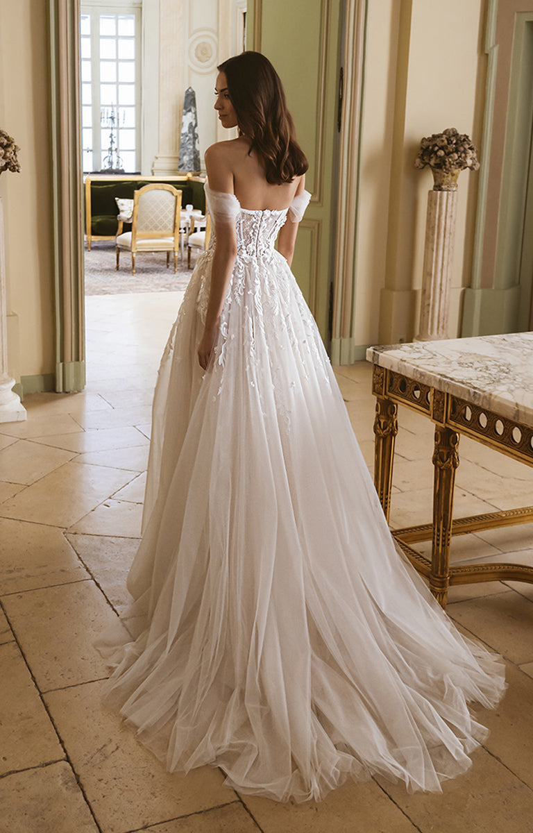 Woman in a white wedding dress standing in an elegant room with high ceilings and large windows.