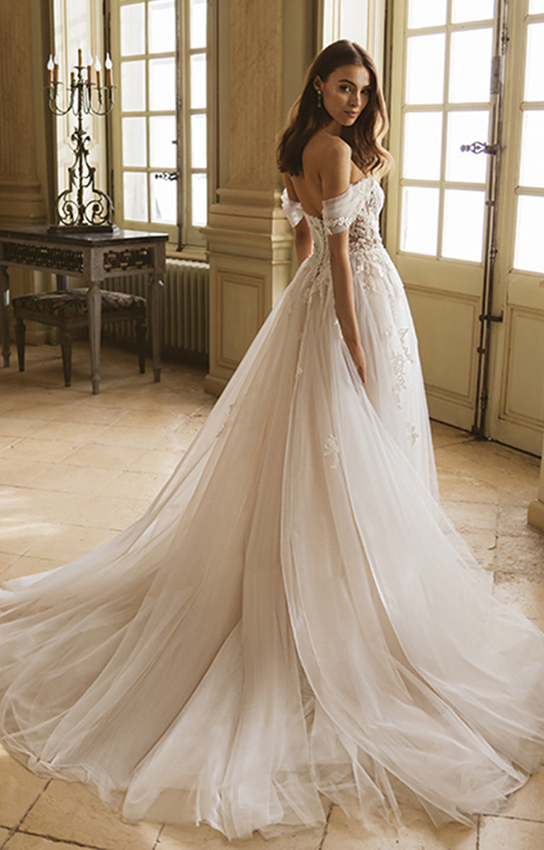 Woman in a white wedding dress standing in a room with large windows and wooden floors.