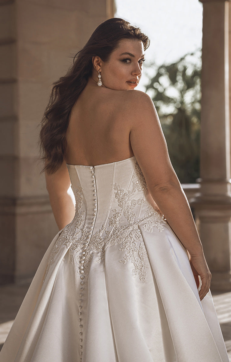 Woman in a white wedding dress with intricate detailing, standing outdoors.