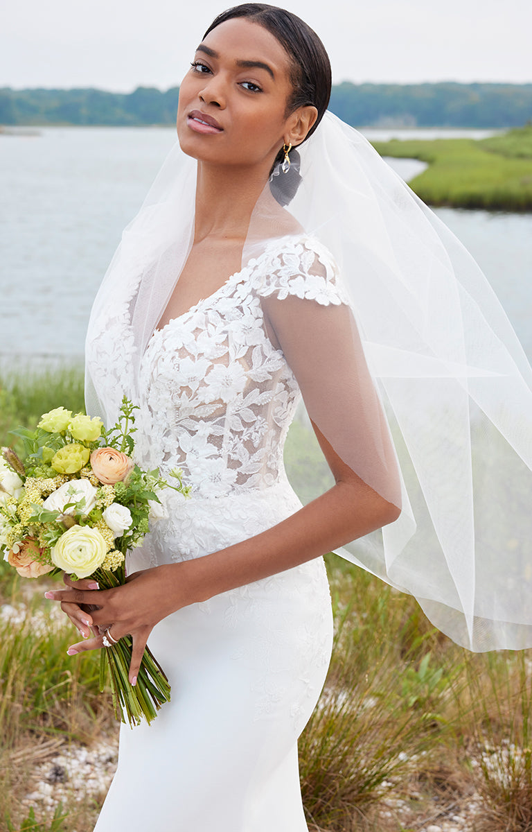 Woman in a white wedding dress with a veil holding flowers by a body of water.