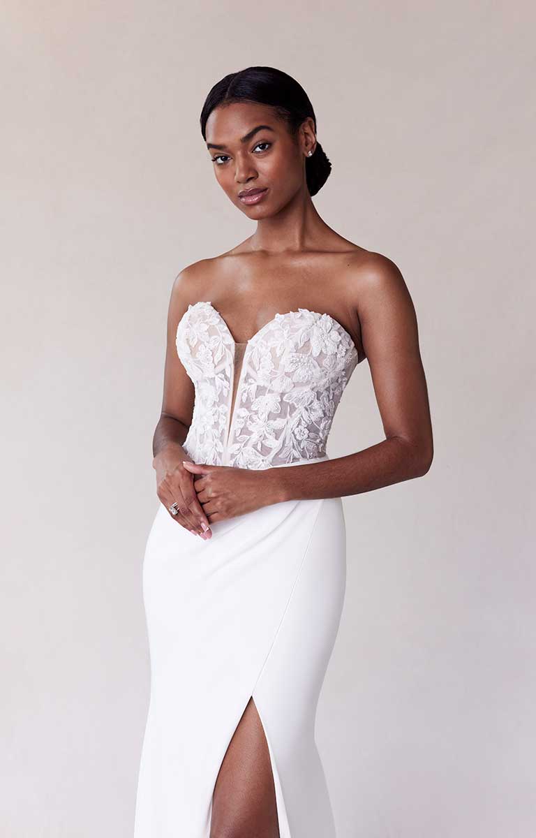 Woman wearing a strapless white lace wedding dress with a slit on a plain background