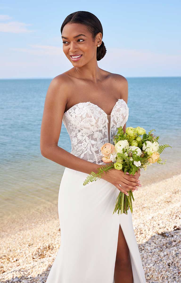 Woman in a white strapless wedding dress holding a bouquet on a beach.