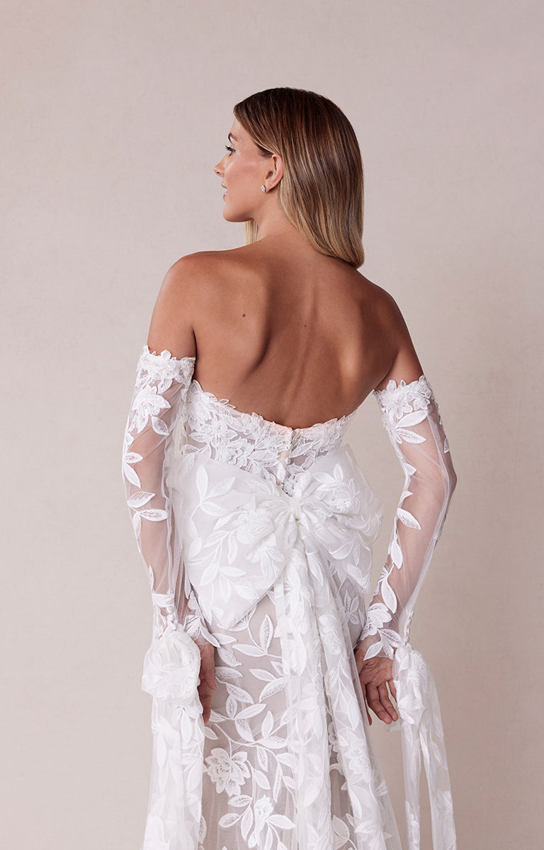 Woman wearing a white lace wedding dress with floral details on a plain background