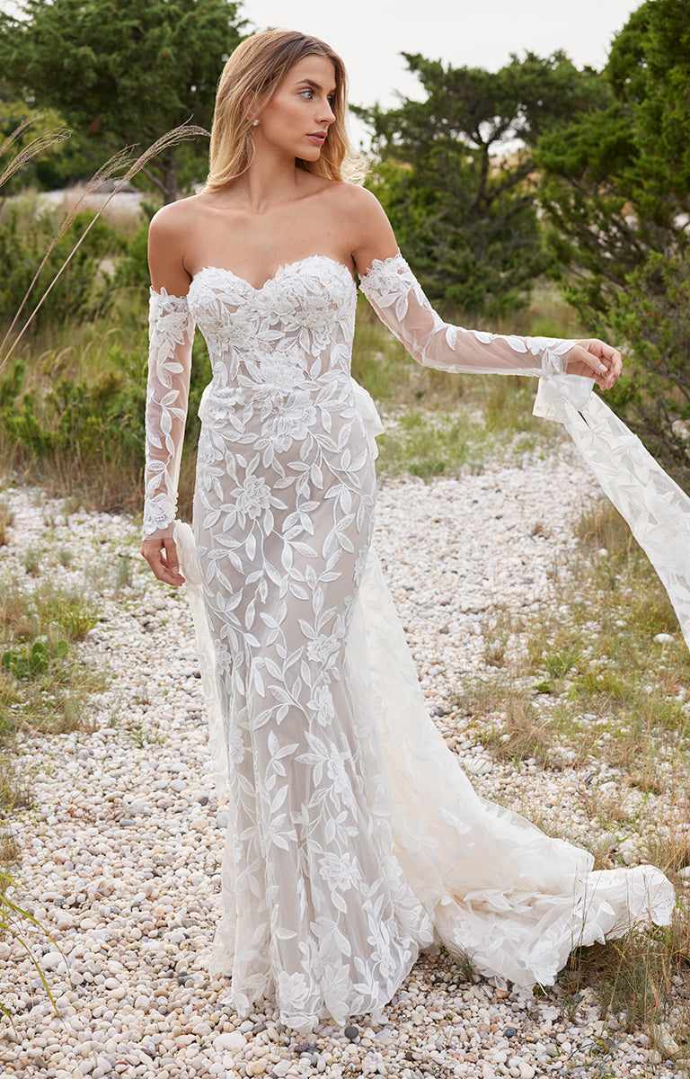 Woman in a white lace wedding dress standing in a natural setting with trees and gravel.