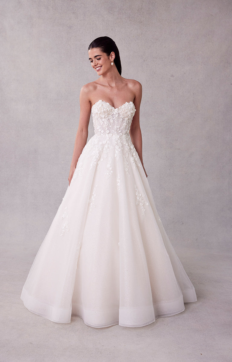 Woman wearing a strapless white wedding dress with floral details against a plain background