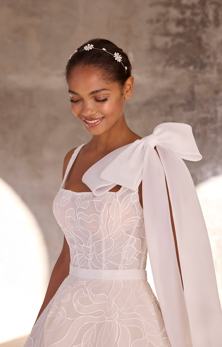 Woman wearing a white wedding dress with a large bow on her shoulder against a neutral background