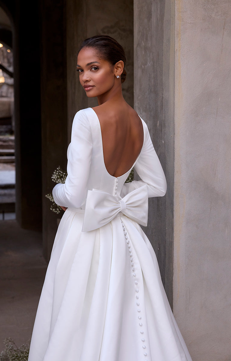 Woman in a white wedding dress with a large bow at the back, standing against a stone wall.