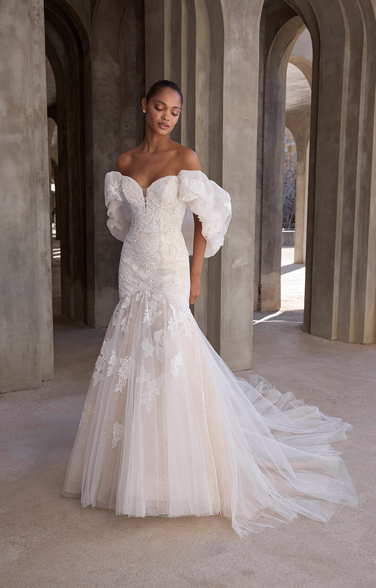 Woman in a white wedding dress with puffed sleeves standing in a sunlit archway.
