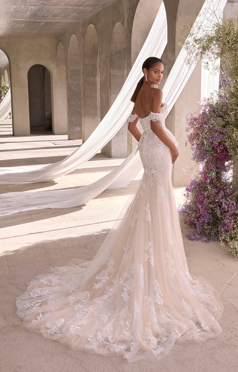 Woman in a white wedding dress with a long train standing in a sunlit outdoor setting.