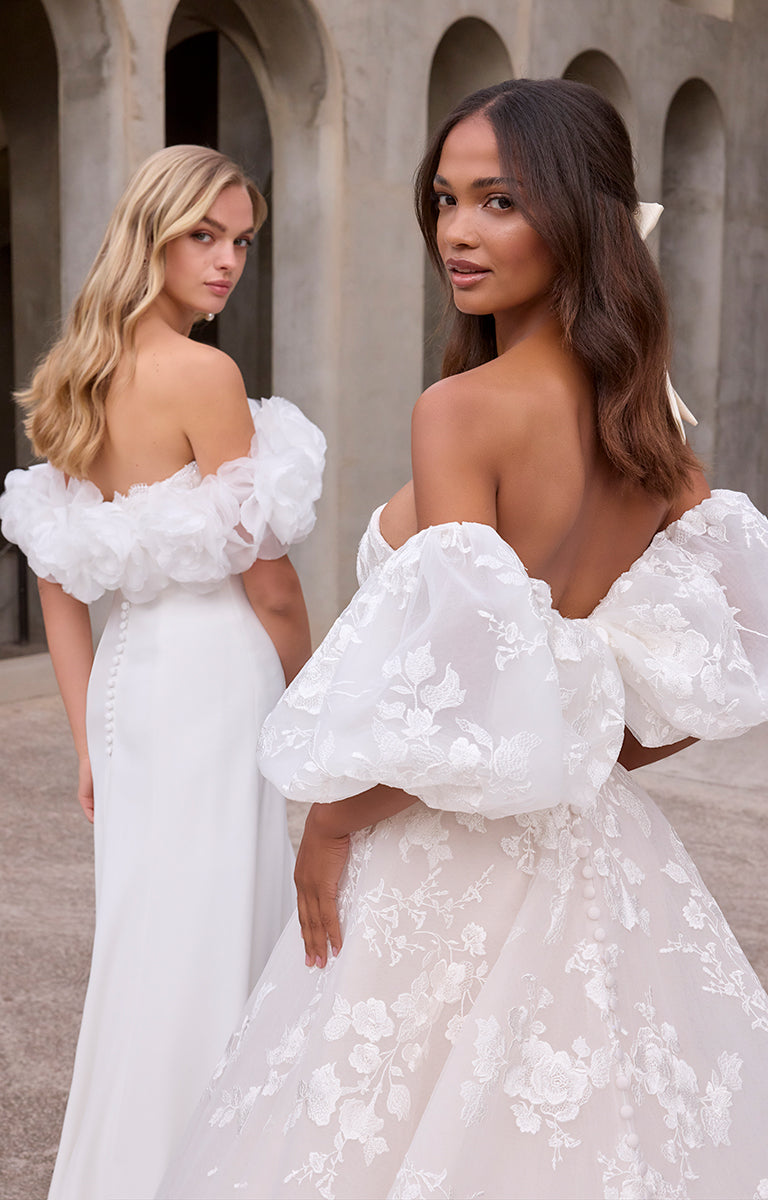 Two women wearing white wedding dresses with floral details in an outdoor setting.
