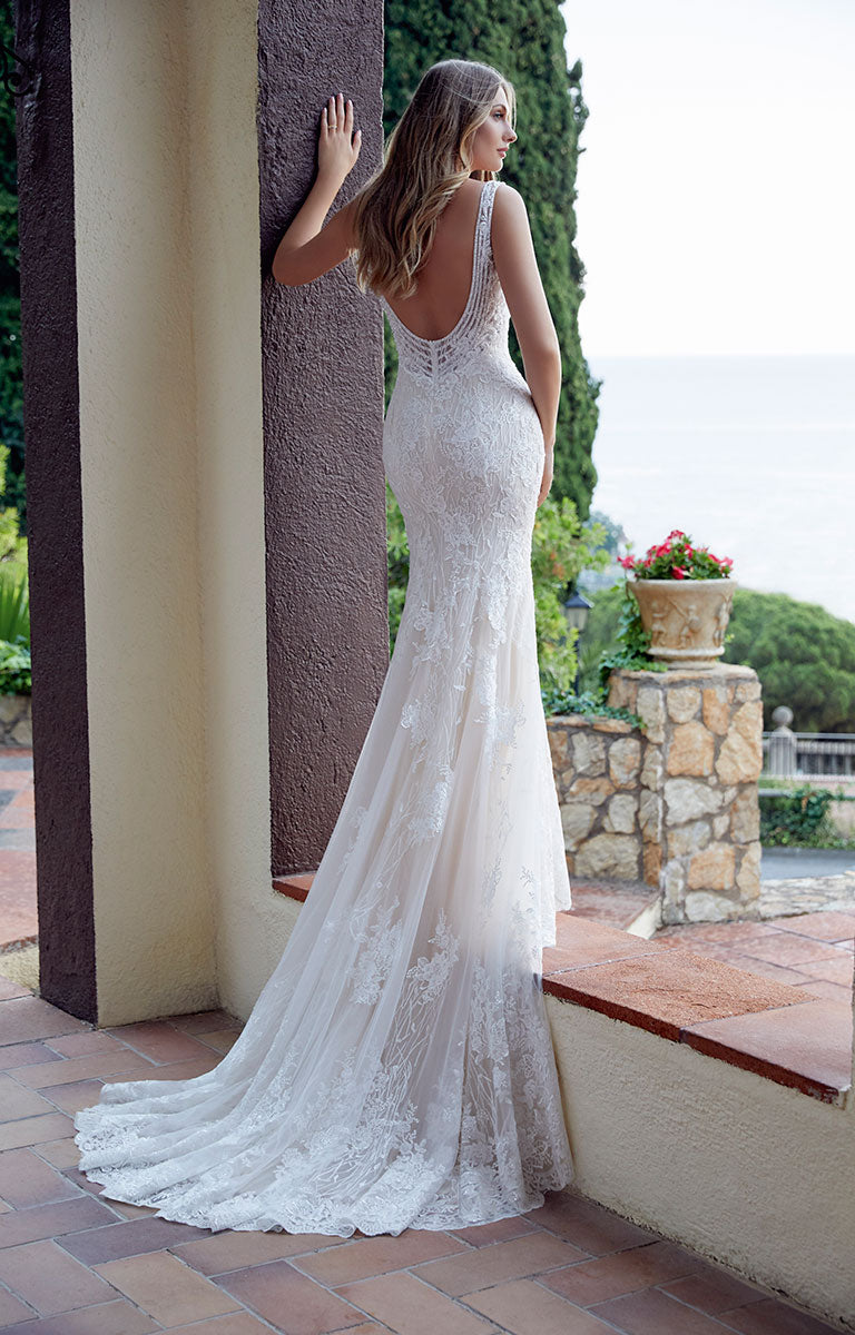 Woman in Dianora 69511 by Veni Infantino - Beaded Fit and Flare Wedding Dress with V-Neckline standing outdoors with greenery and a stone wall in the background.