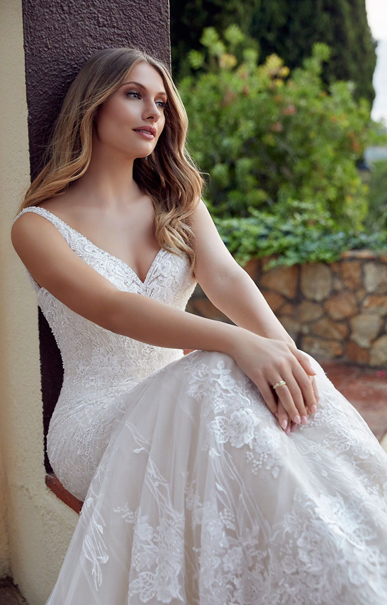 Woman in Dianora 69511 by Veni Infantino - Beaded Fit and Flare Wedding Dress with V-Neckline sitting outdoors with greenery in the background