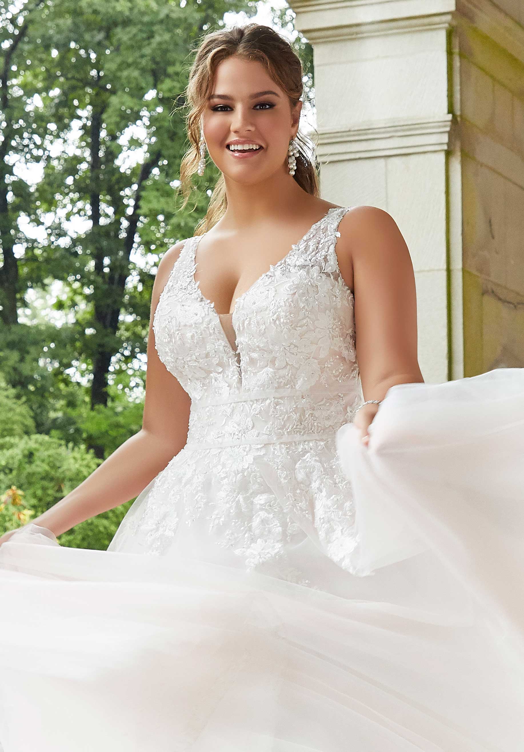 Woman in a white wedding dress standing outdoors with greenery and a building in the background