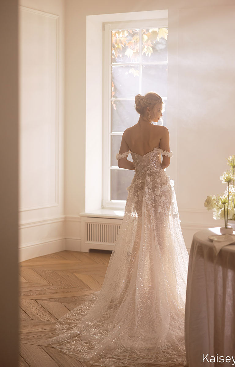Woman in a white wedding dress standing in a softly lit room with a window.