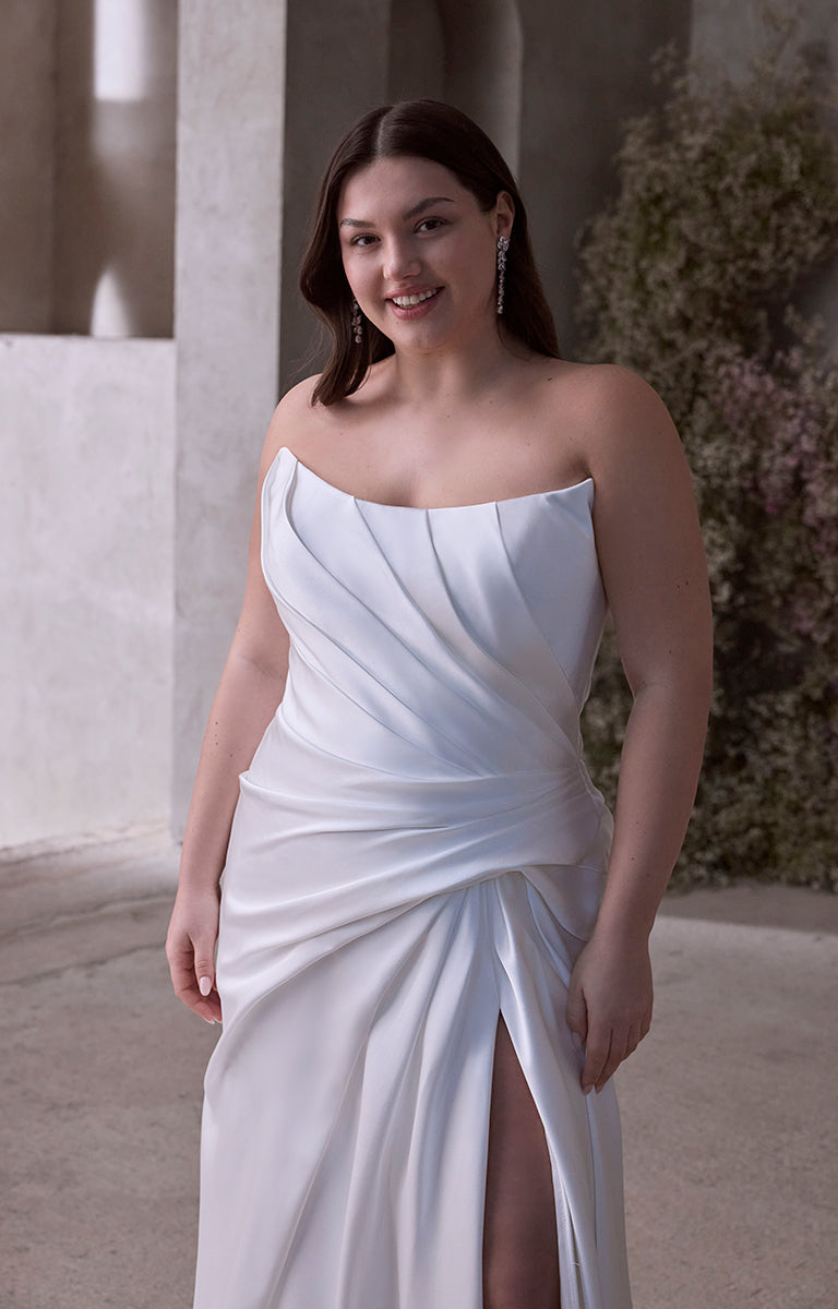 Woman wearing Vipsania by Morilee Julietta - a white strapless gown with a slit, standing outdoors.