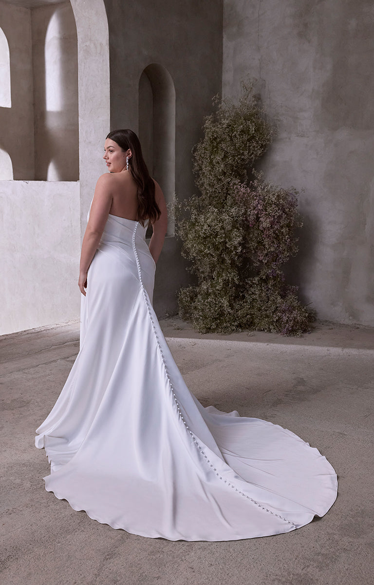 Woman in Vipsania by Morilee Julietta - a white wedding dress standing in a sunlit outdoor setting with arches and plants.