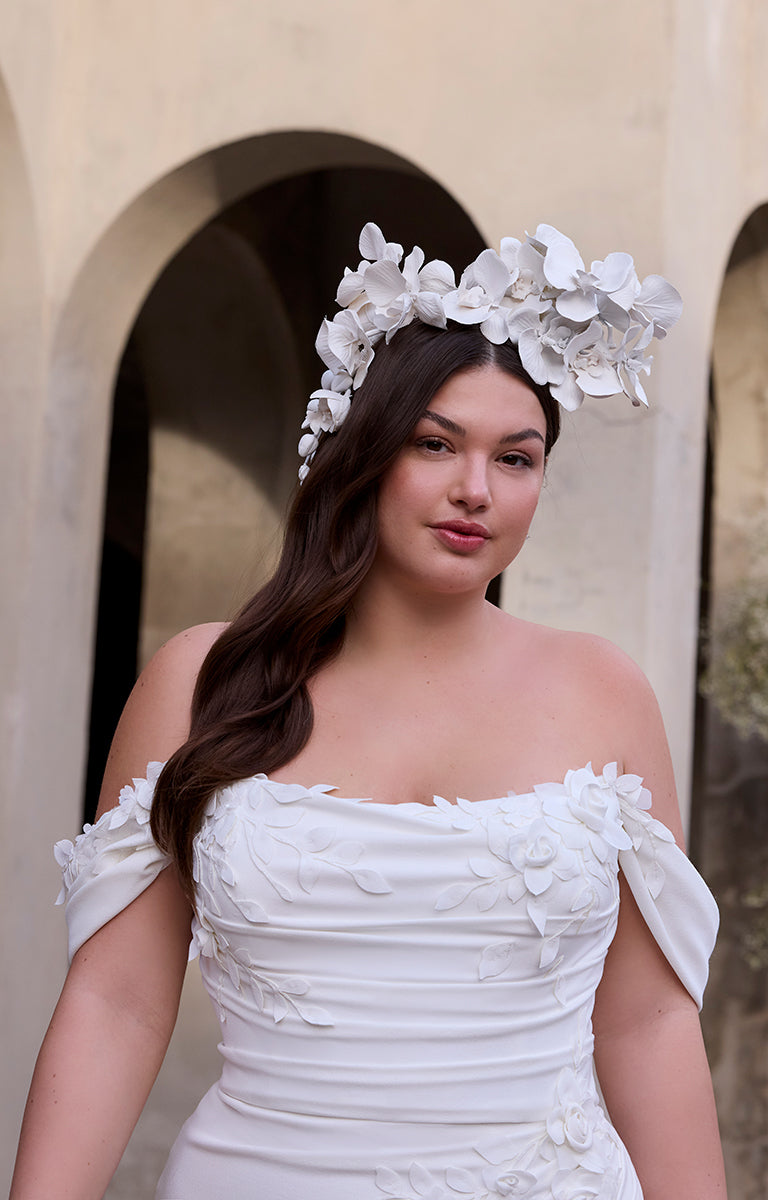 Woman wearing Rea by Morilee Julietta - a white floral headpiece and off-shoulder wedding dress in front of a stone archway.