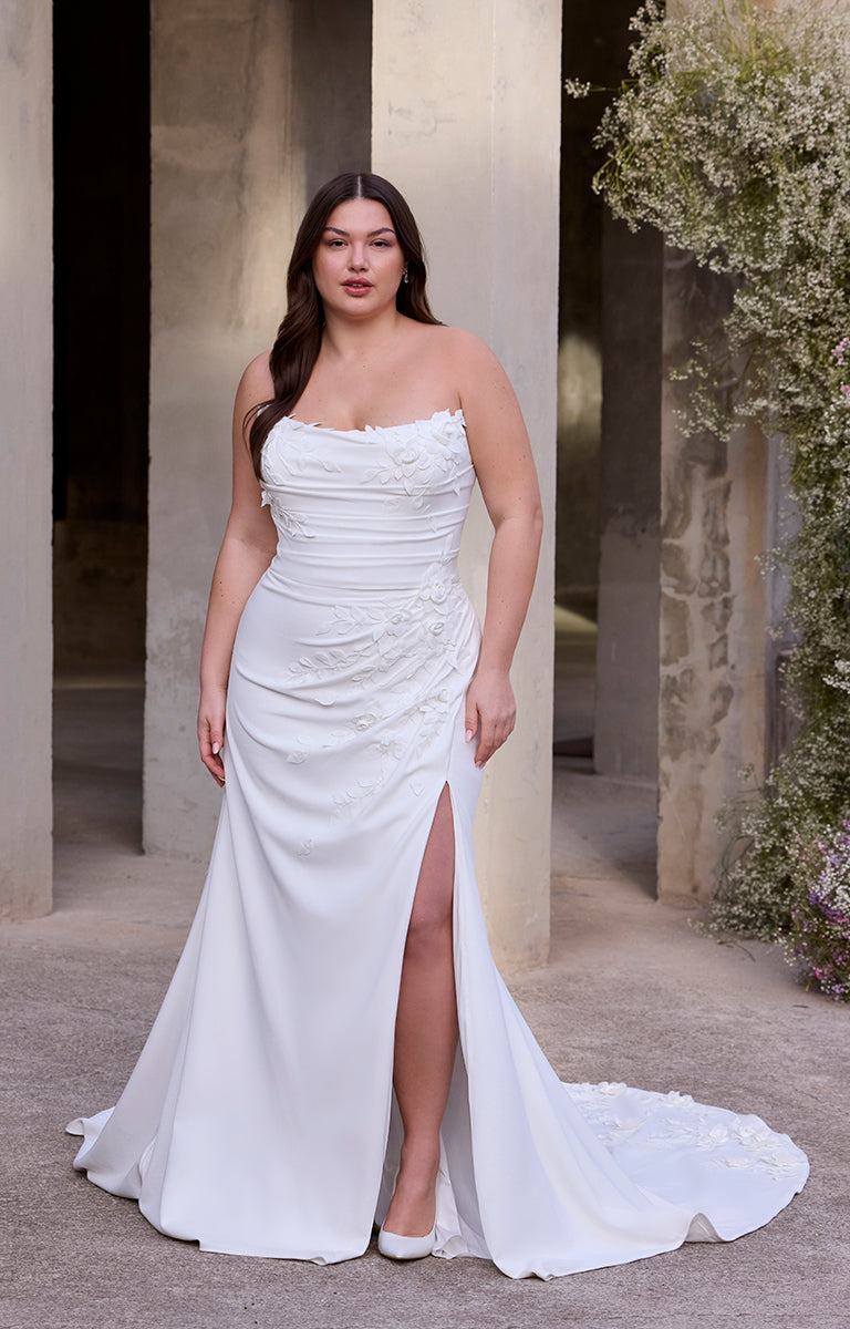 Woman in Rea by Morilee Julietta - a white strapless gown with a slit standing in an outdoor setting.