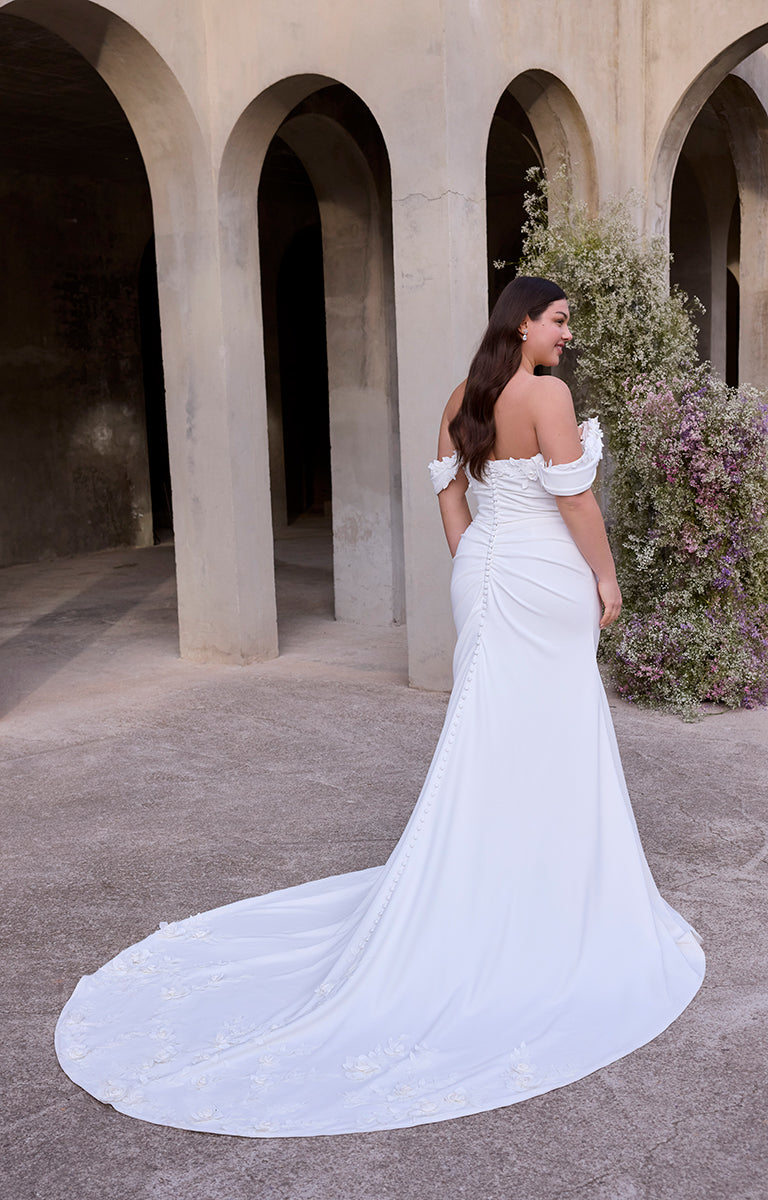 Woman in Rea by Morilee Julietta - a white off-shoulder wedding dress standing in an archway with floral decorations.