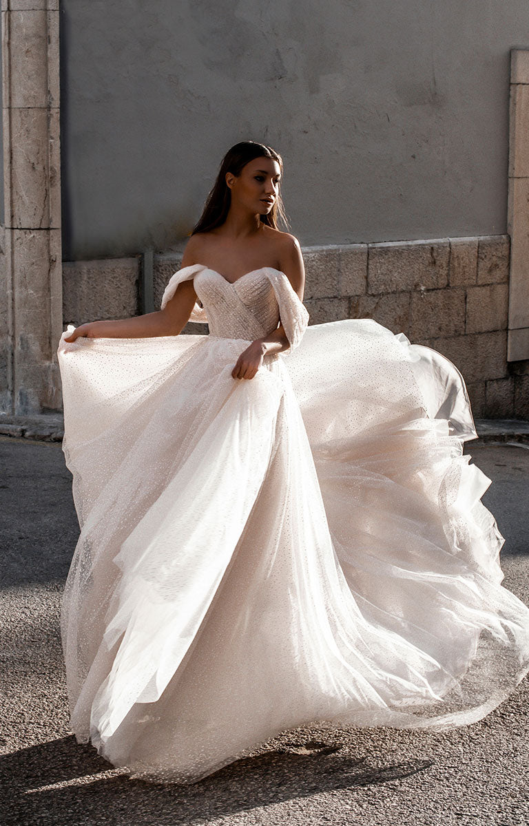 Woman in Gita-Marie E176 by Abella - Dotted Tulle A-Line Wedding Dress with Off-Shoulder Sleeves standing outdoors against a stone wall.