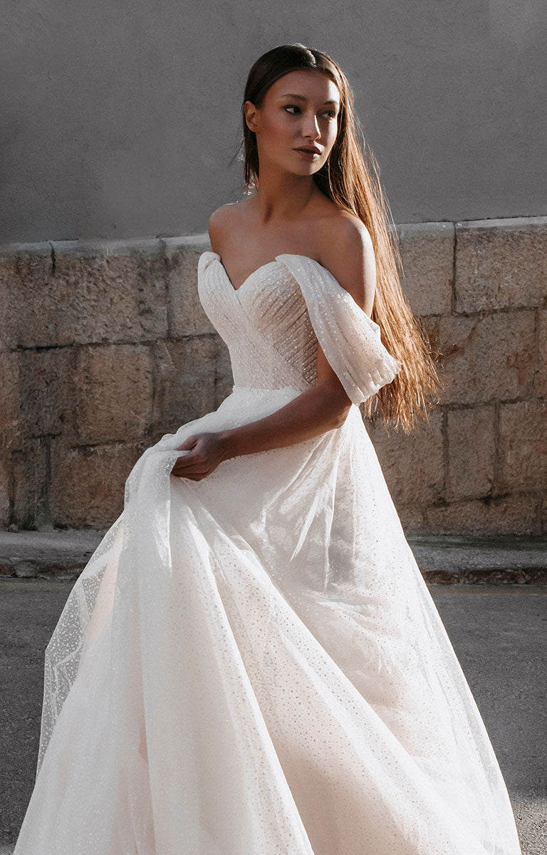 Woman wearing Gita-Marie E176 by Abella - Dotted Tulle A-Line Wedding Dress with Off-Shoulder Sleeves standing against a stone wall.