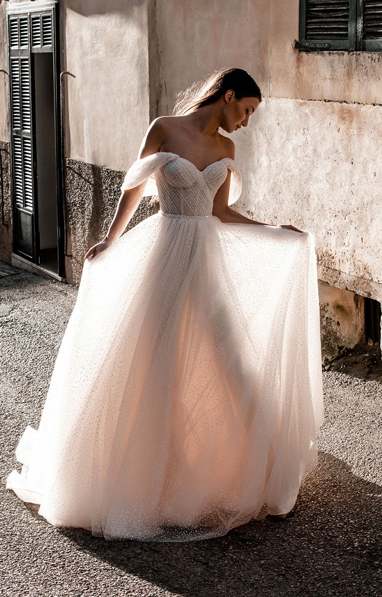 Woman in Gita-Marie E176 by Abella - Dotted Tulle A-Line Wedding Dress with Off-Shoulder Sleeves standing in front of a textured wall.