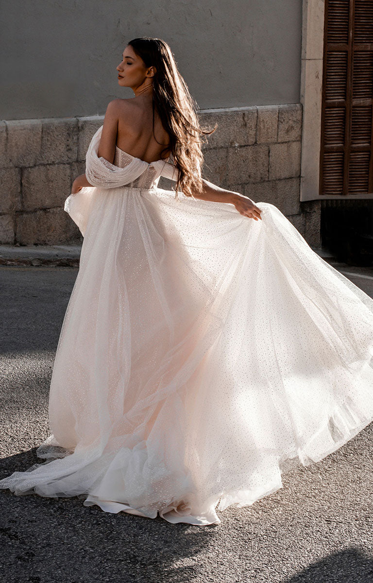 Woman in Gita-Marie E176 by Abella - Dotted Tulle A-Line Wedding Dress with Off-Shoulder Sleeves standing on a street.