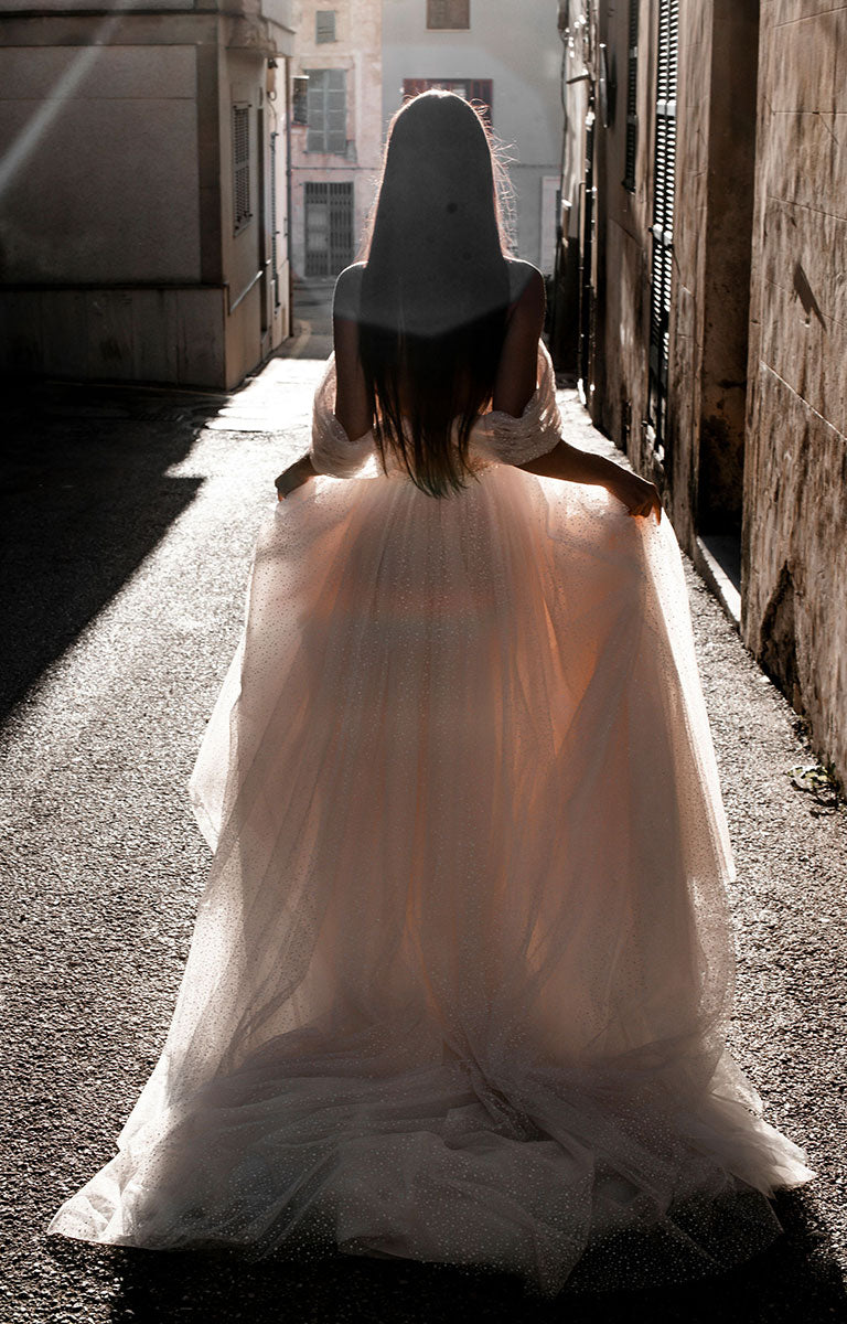 Woman in Gita-Marie E176 by Abella - Dotted Tulle A-Line Wedding Dress with Off-Shoulder Sleeves walking away down a sunlit alleyway.