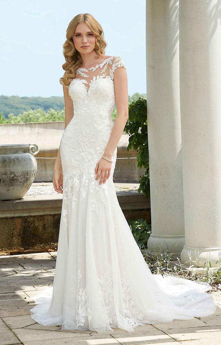 Woman in Daisy Wedding Dress by Morilee - Style 5946 – Botanical Lace Mermaid Gown with Illusion Neckline standing outdoors with columns and greenery in the background