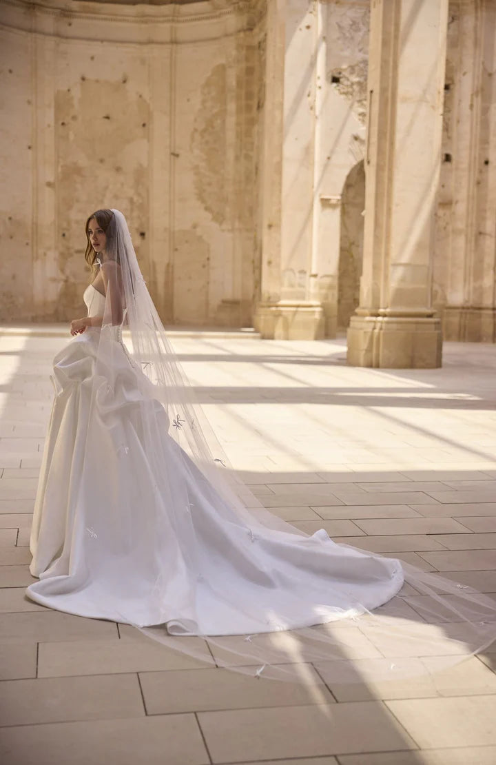 Bride in a white wedding dress with a long veil standing in a sunlit stone hallway.