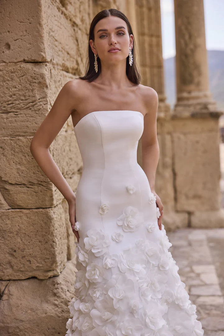 Woman in a white strapless wedding dress with floral details standing against a stone wall.