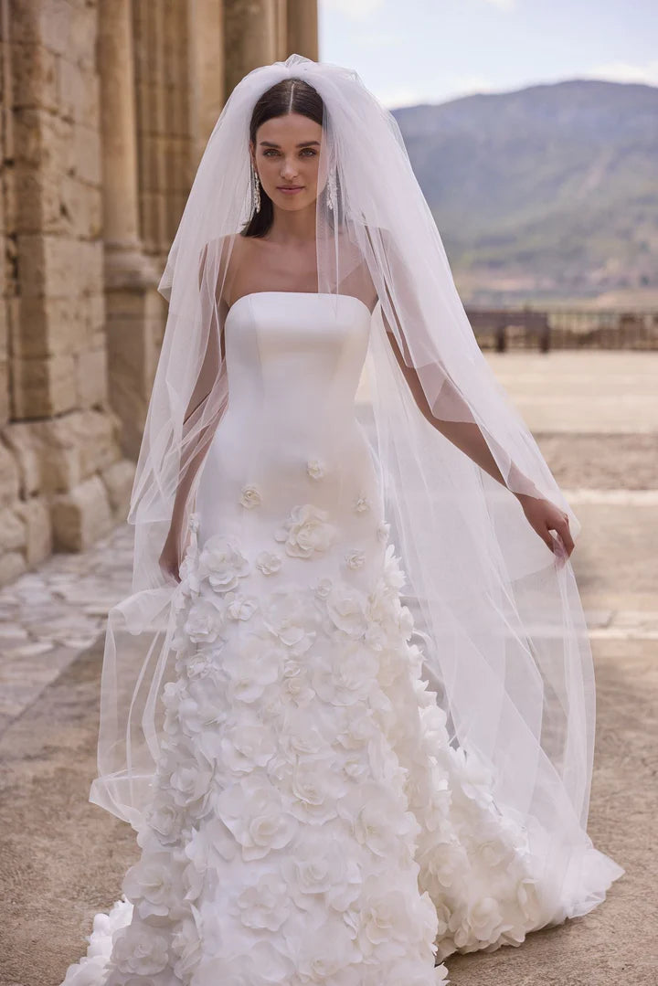 Woman in a white wedding dress with floral details and a long veil outdoors.