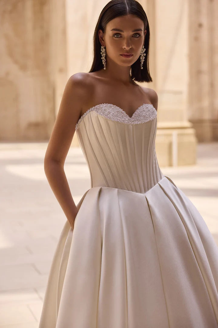 Woman wearing a strapless wedding dress with beaded top and pleated skirt in an outdoor setting.