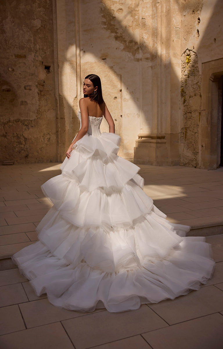 Woman in a white wedding dress with a ruffled skirt standing in a sunlit stone hallway.