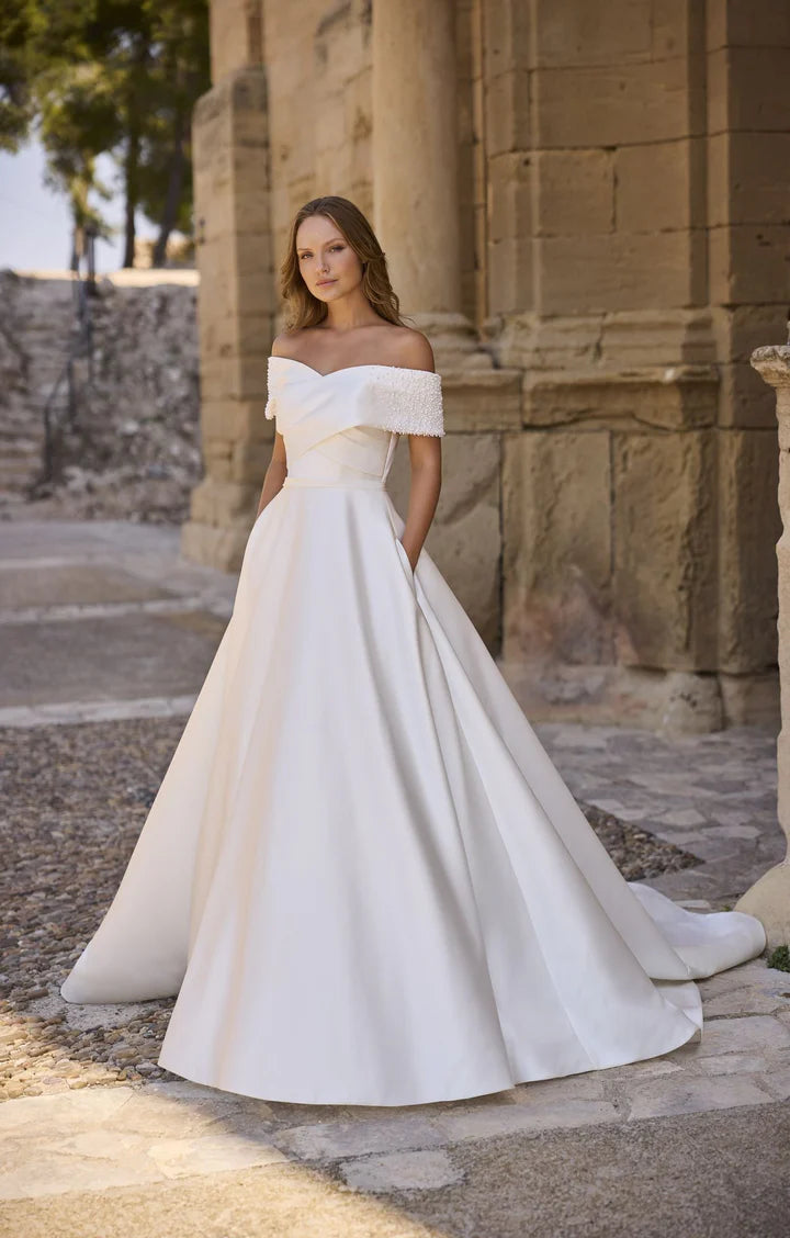 Woman in a white off-shoulder wedding dress standing in front of ancient stone architecture.