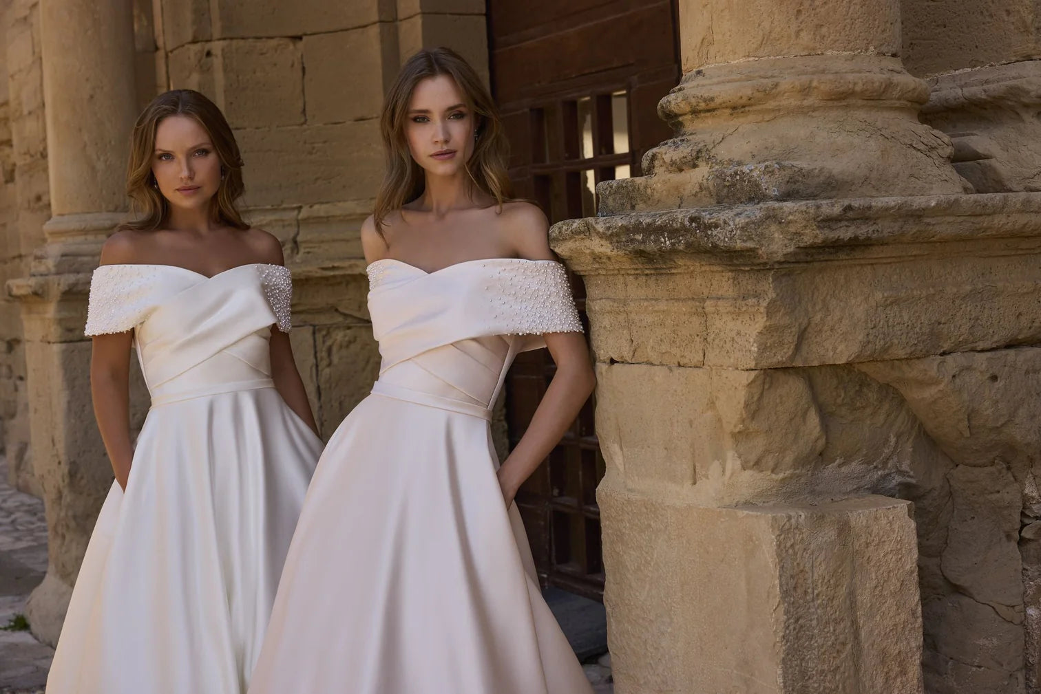 Two women in white off-shoulder dresses standing in front of stone architectural elements.
