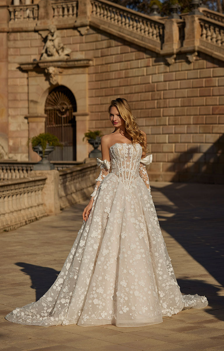 Woman in 63113 Hildagarde by Veni Infantino Bridal - Floral Lace Ball Gown with Corset Bodice and Detachable Bow Sleeves standing in front of an elegant building.