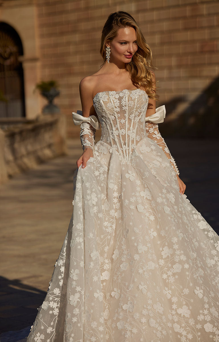 Woman in 63113 Hildagarde by Veni Infantino Bridal - Floral Lace Ball Gown with Corset Bodice and Detachable Bow Sleeves standing outdoors.