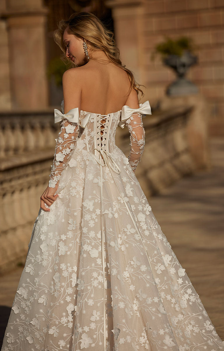 Woman wearing 63113 Hildagarde by Veni Infantino Bridal - Floral Lace Ball Gown with Corset Bodice and Detachable Bow Sleeves with floral patterns in an outdoor setting.