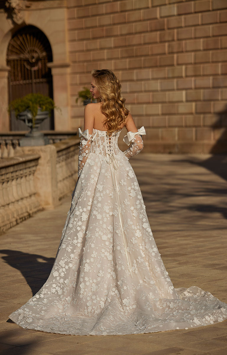 Woman in 63113 Hildagarde by Veni Infantino Bridal - Floral Lace Ball Gown with Corset Bodice and Detachable Bow Sleeves standing in front of a stone building.