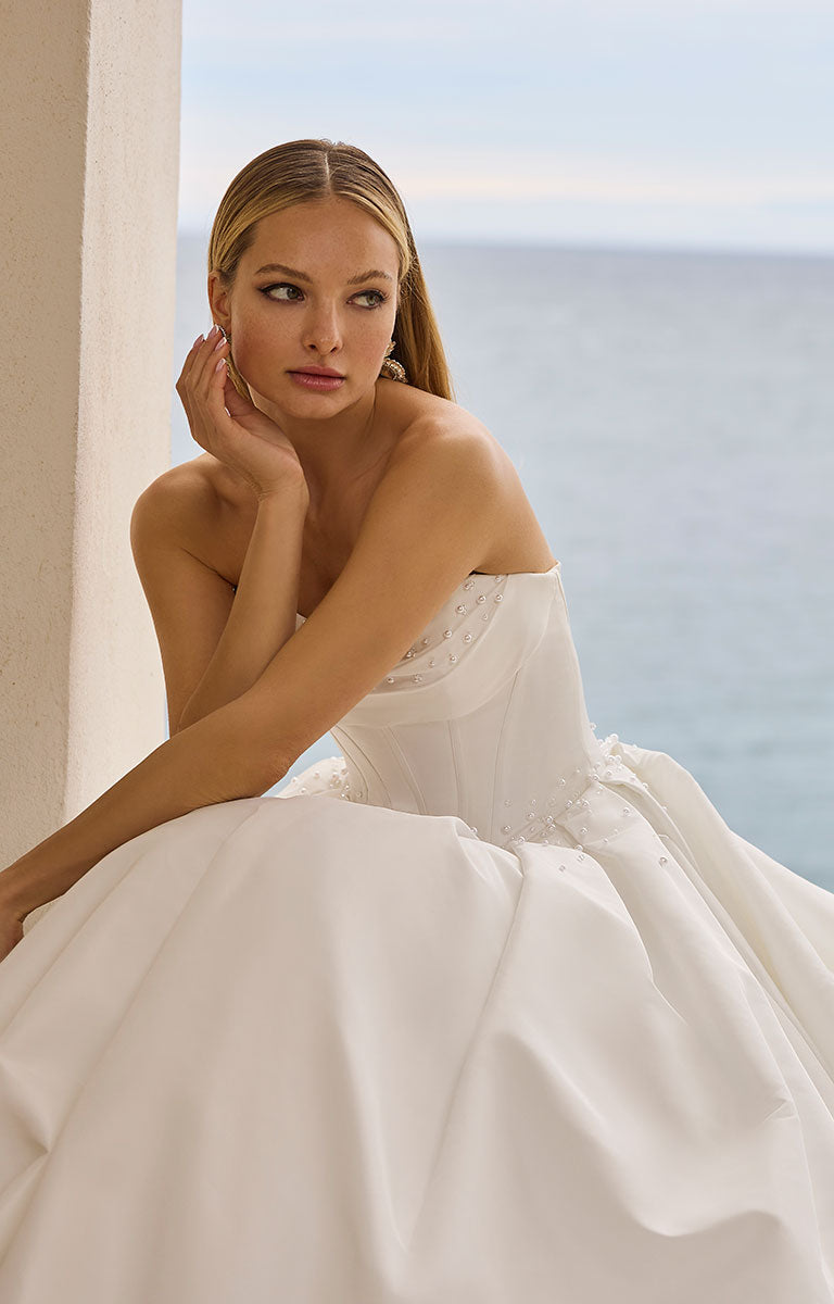 Woman in 63125 Hania by Veni Infantino Bridal - Strapless Taffeta Ball Gown with Beaded Bodice sitting by a window with a scenic view.