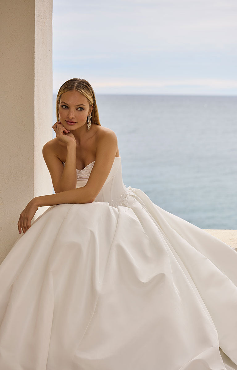 Woman in 63125 Hania by Veni Infantino Bridal - Strapless Taffeta Ball Gown with Beaded Bodice standing by a window with a scenic view of the ocean.