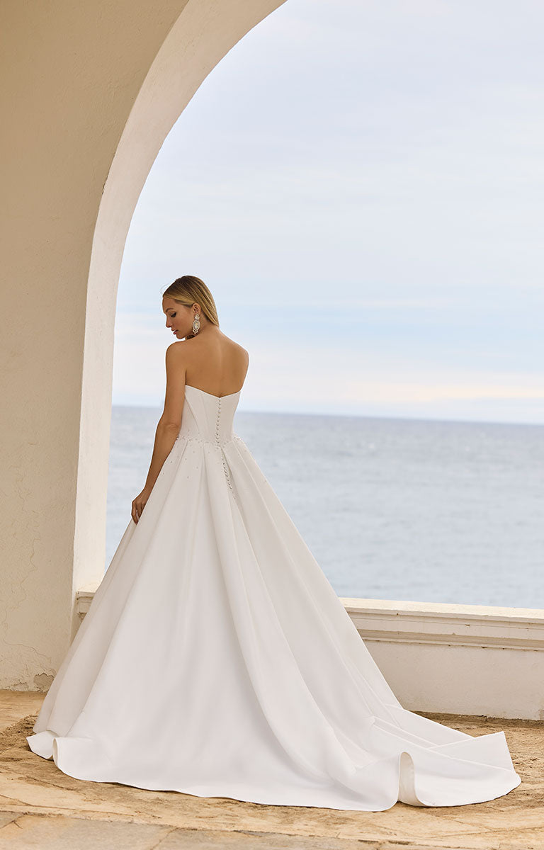 Woman in 63125 Hania by Veni Infantino Bridal - Strapless Taffeta Ball Gown with Beaded Bodice standing by a large archway with a scenic view of water.